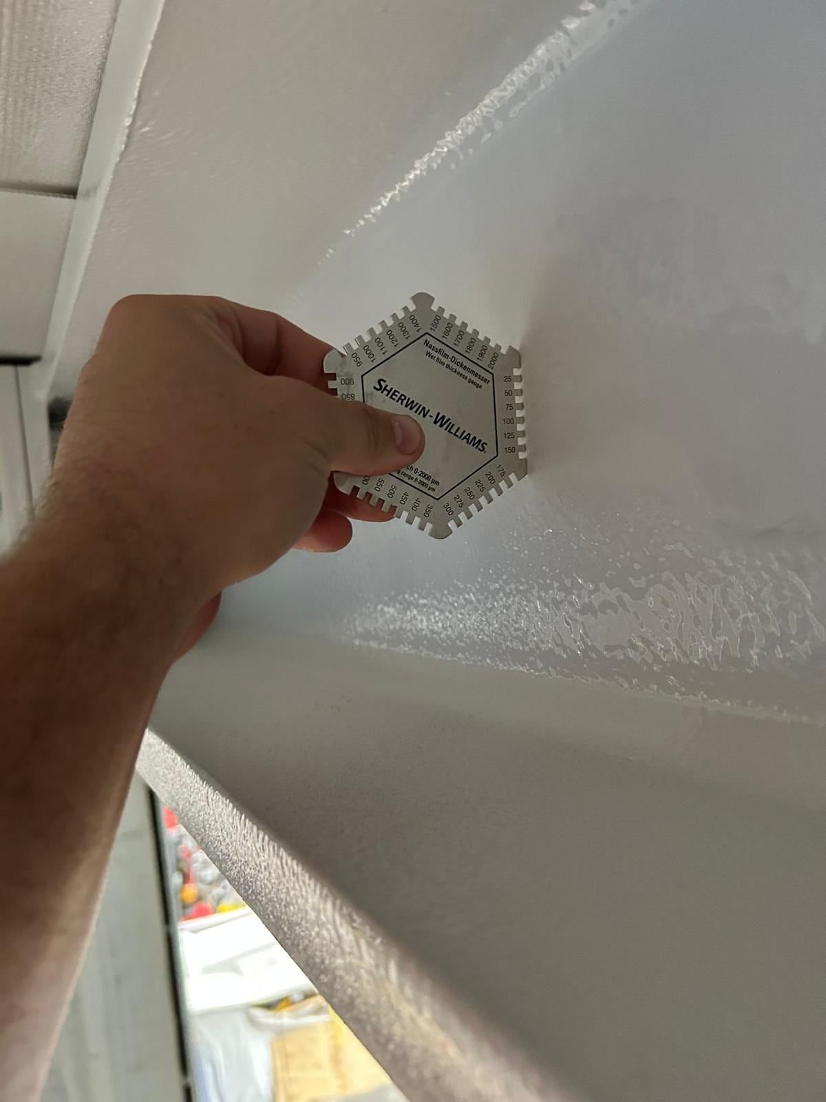 Hand holding a hexagonal wet film gauge against freshly applied white paint on a steel section